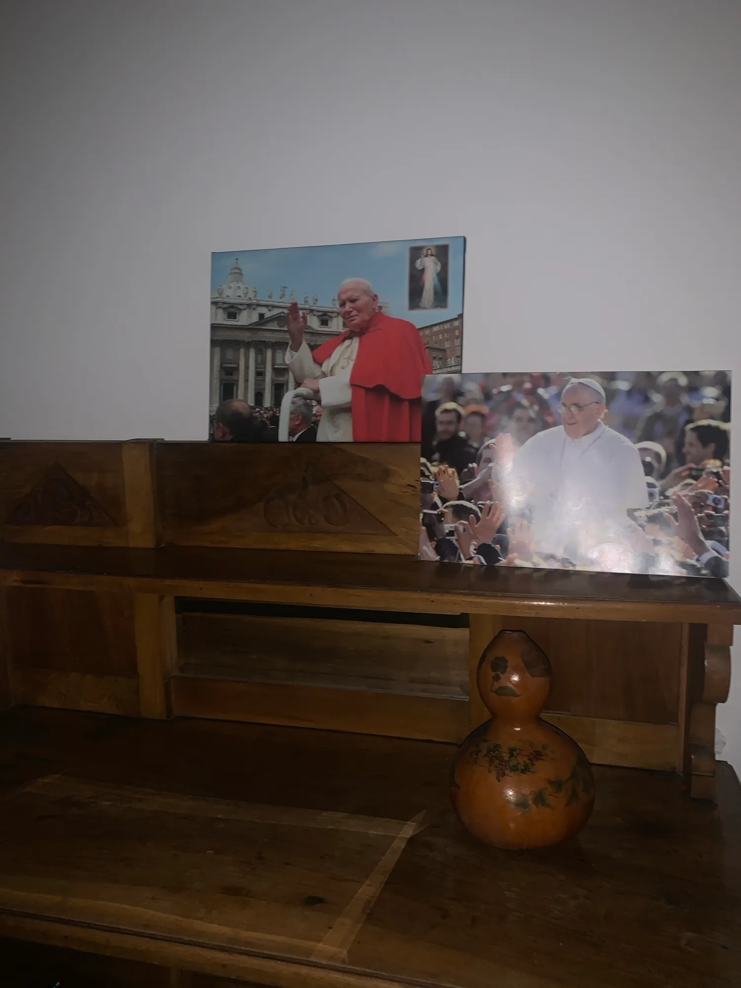 Zwei gerahmte Papstfotos auf einer Holzbank, davor ein bemalter Flaschenkürbis