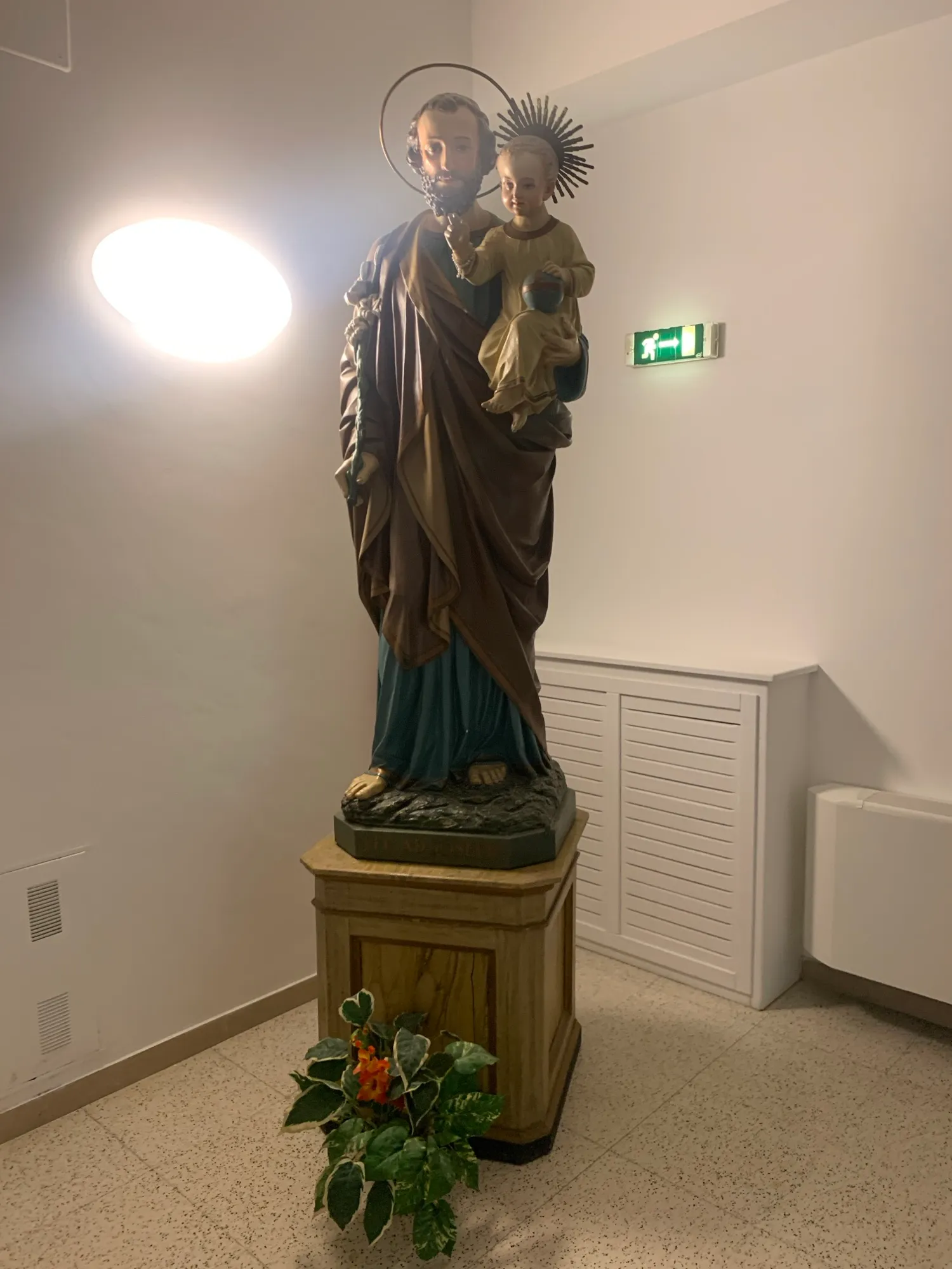 Statue des heiligen Josef mit dem Jesuskind auf einem Sockel, davor Blumen, daneben ein Notausgangsschild